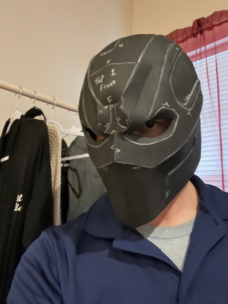 May include: A black, angular mask with white markings and the text "Top 1 Front" is worn by a person. The mask covers the entire face and has eye and mouth openings. The mask appears to be a prototype or a work in progress.