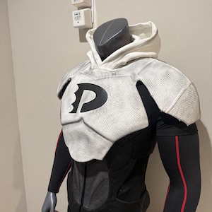 Può includere: Un costume su un manichino con un top con cappuccio bianco e un'armatura per le spalle con un logo "P" nero. Il costume ha maniche nere con finiture rosse e una sezione del busto nera. Tema generale del supereroe.