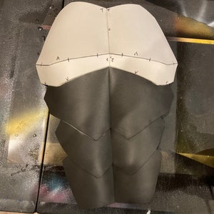May include: A white and black paper pattern for a costume, with markings for cutting and sewing. The pattern is for a chest piece with a series of overlapping scales.