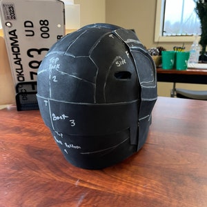 May include: A black, segmented helmet prototype with white markings and the words "Top Back", "Side", and "Back" written on it. The helmet is on a wooden surface. The Oklahoma license plate is visible in the background.