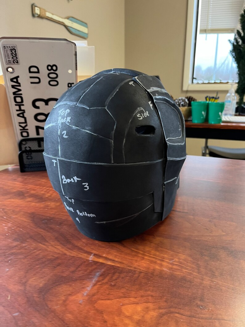 May include: A black, segmented helmet prototype with white markings and the words "Top Back", "Side", and "Back" written on it. The helmet is on a wooden surface. The Oklahoma license plate is visible in the background.