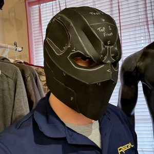 May include: Black helmet with eye cutouts and white markings, labeled "Top 1 Front". The helmet is worn by a person wearing a blue shirt with the word "REGA" in gold lettering. The helmet appears to be a cosplay or costume piece.