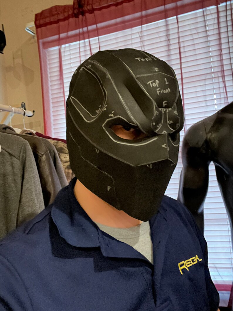 May include: Black helmet with eye cutouts and white markings, labeled "Top 1 Front". The helmet is worn by a person wearing a blue shirt with the word "REGA" in gold lettering. The helmet appears to be a cosplay or costume piece.