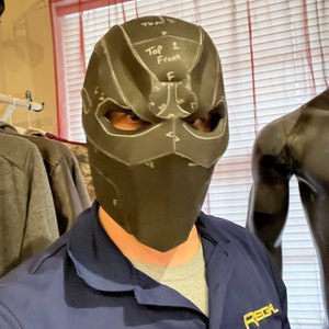 May include: A black mask with white markings and the text "Top 1 Front" is worn by a person. The mask has eye openings and a geometric design. The person is wearing a blue shirt with the word "REGA" printed on it.