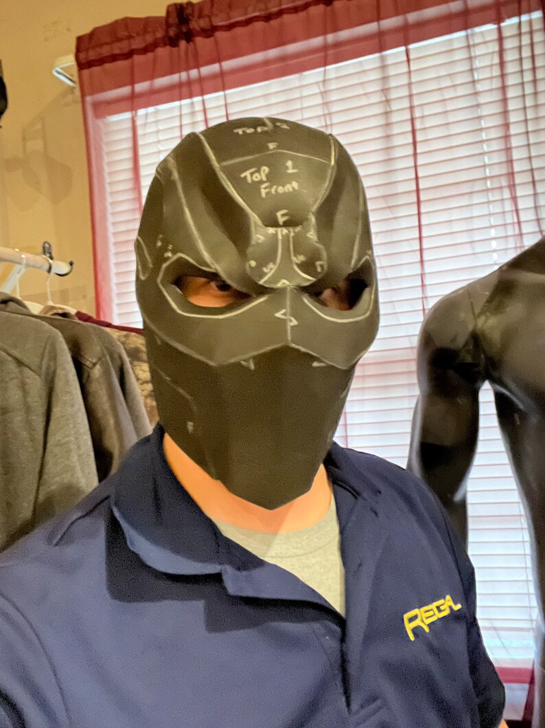 May include: A black mask with white markings and the text "Top 1 Front" is worn by a person. The mask has eye openings and a geometric design. The person is wearing a blue shirt with the word "REGA" printed on it.