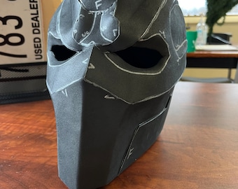 May include: A black, angular mask with cutouts for the eyes and mouth. White lines and text, including "Top 1 Front F", are drawn on the mask's surface, indicating a pattern or design. The mask is positioned on a wooden surface.