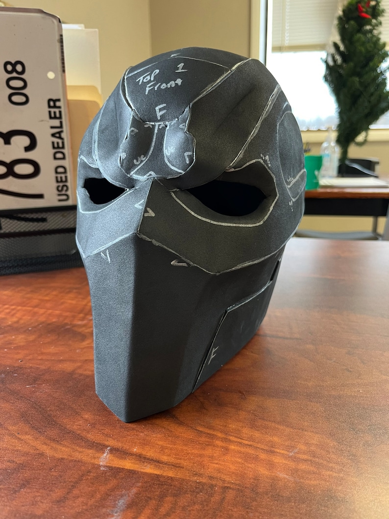 May include: A black, angular mask with cutouts for the eyes and mouth. White lines and text, including "Top 1 Front F", are drawn on the mask's surface, indicating a pattern or design. The mask is positioned on a wooden surface.