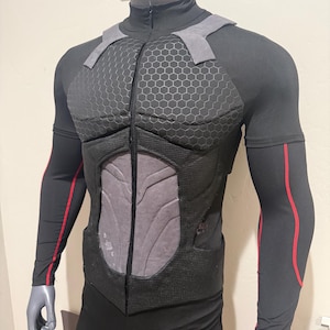 May include: Black and gray tactical bodysuit with a high collar and zipper closure. The chest and shoulders feature padded armor with a honeycomb pattern. Red accents run down the sleeves. The suit is displayed on a mannequin.