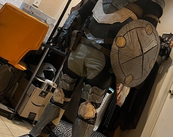 May include: A person wearing a full-body costume of a fantasy warrior, with a helmet, armor, and a shield. The costume is made of dark gray and silver materials, and the shield is decorated with a circular design.