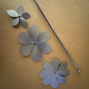 Op de afbeelding: Drie zilveren metalen bloemblaadjes en een lange zilveren metalen staaf zijn gerangschikt op een lichtbruin oppervlak. De bloemblaadjes hebben een eenvoudig ontwerp met een gat in het midden. De staaf heeft een kleine bal aan het uiteinde.