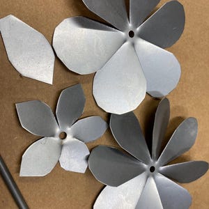 May include: Close-up of several unfinished metal flower petals and assembled flowers. The petals are silver and have a matte finish. The flowers have a hole in the center, and the petals are slightly curved. The background is a light brown.
