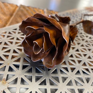May include: A handcrafted metal rose sculpture with a detailed, layered petal design. The rose is a warm, rusty brown colour, with a stem and leaves. The sculpture is placed on a metal surface with a geometric pattern.