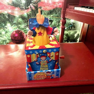 May include: A red and blue toy jack-in-the-box with a clown popping out. The clown is wearing a yellow hat and a blue and red costume. The box has a yellow and red striped design and says "Silly Circus" on the front.
