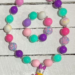 Pu&ograve; includere: Un colorato set di collana e braccialetto con grandi perline rotonde bianche, rosa, viola e verde acqua. La collana include una statuina di Paperina con un gelato. Le perline sono infilate su una catena color argento.