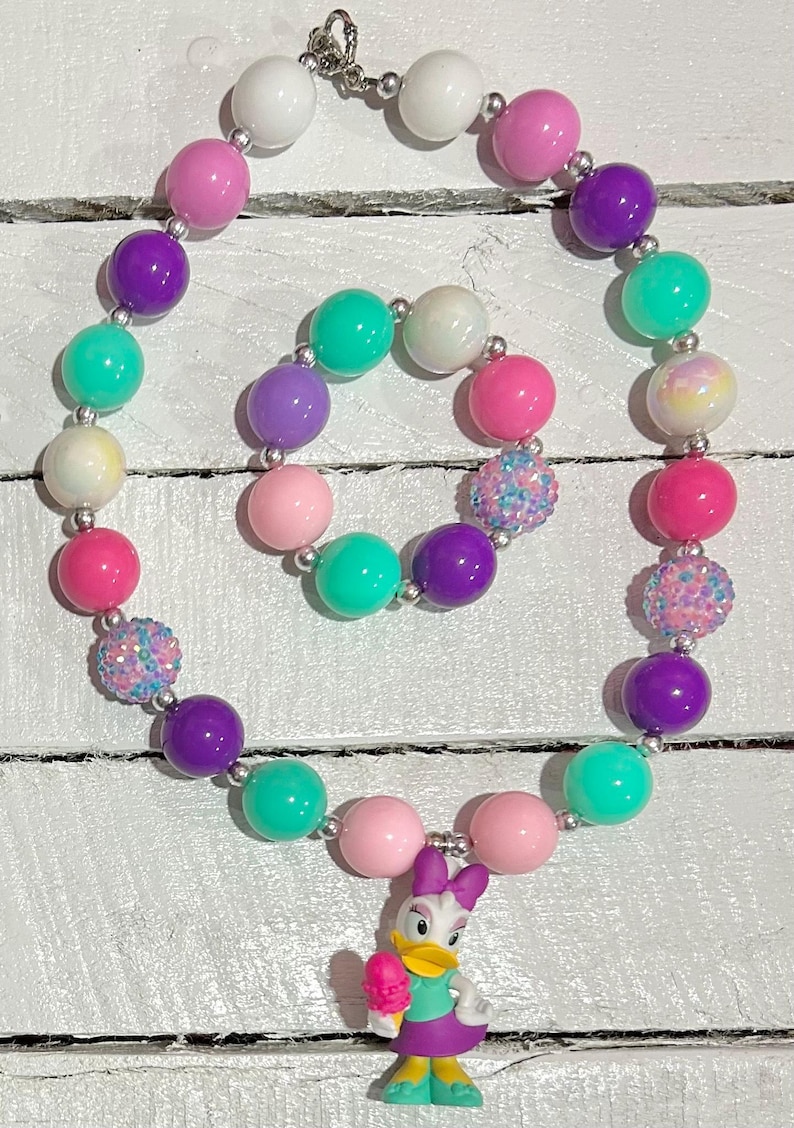 Pu&ograve; includere: Un colorato set di collana e braccialetto con grandi perline rotonde bianche, rosa, viola e verde acqua. La collana include una statuina di Paperina con un gelato. Le perline sono infilate su una catena color argento.