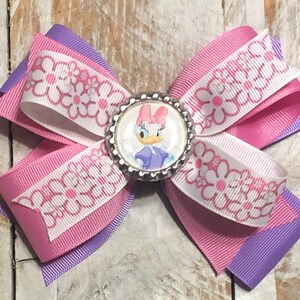 Pu&ograve; includere: Un fiocco per capelli fatto a mano con un'immagine di Daisy Duck al centro. Il fiocco &egrave; realizzato con nastri a strati in rosa, bianco con un motivo a margherite e viola. Un accessorio ideale per aggiungere un tocco di magia Disney.