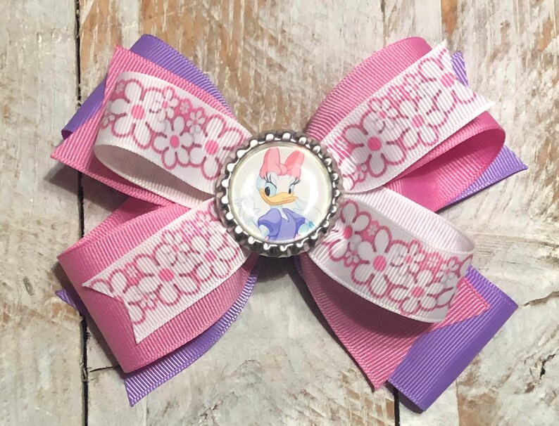 Pu&ograve; includere: Un fiocco per capelli fatto a mano con un'immagine di Daisy Duck al centro. Il fiocco &egrave; realizzato con nastri a strati in rosa, bianco con un motivo a margherite e viola. Un accessorio ideale per aggiungere un tocco di magia Disney.