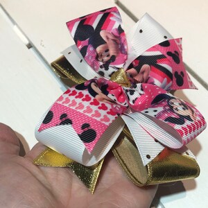 May include: A pink, white, and gold bow with a Minnie Mouse print. The bow is made of ribbon and has a gold glitter finish.