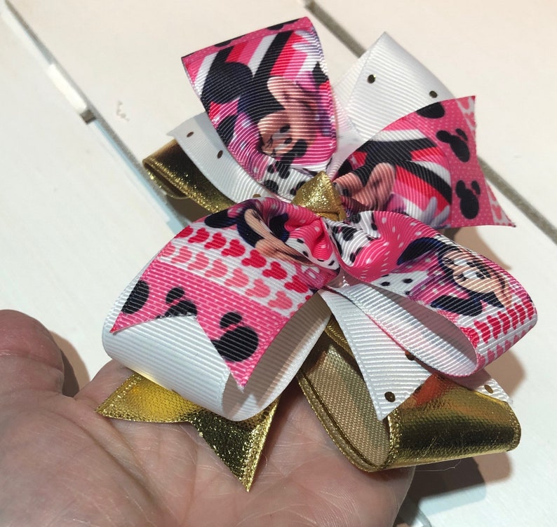 May include: A pink, white, and gold bow with a Minnie Mouse print. The bow is made of ribbon and has a gold glitter finish.