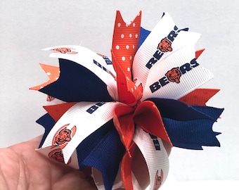 May include: A multi-colored hair bow with white, orange, and blue ribbons. The ribbons have a white background with the word "BEARS" printed in black and a brown bear logo. The bow has a white polka dot ribbon.