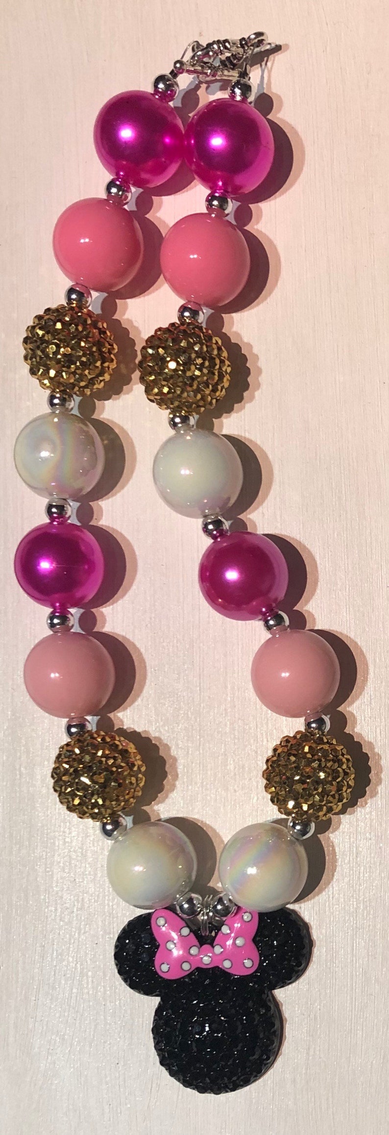 May include: A pink, white, and gold beaded necklace with a black and pink Minnie Mouse charm. The necklace is made with large, round beads and has a silver clasp.