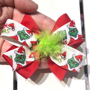 Peut inclure: Un nœud de Noël rouge et blanc Grinch avec un accent de plume verte. Le nœud présente un motif répété du Grinch portant un chapeau de Père Noël et les mots "Grinchmas".