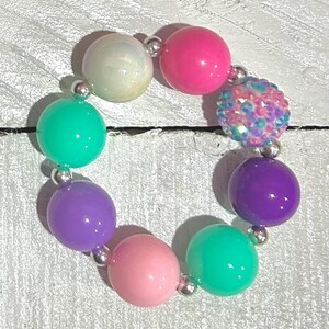 Pu&ograve; includere: Un braccialetto colorato con grandi perline rotonde in vari colori, tra cui rosa, viola, verde e bianco iridescente. Le perline sono collegate da piccoli connettori color argento. Una perlina &egrave; ricoperta di paillettes iridescenti.