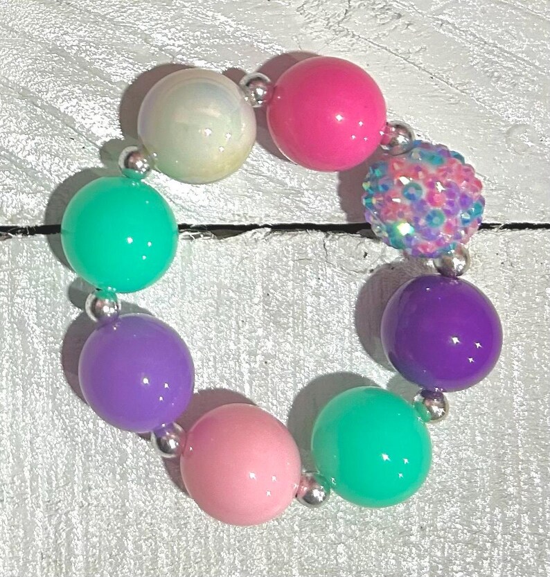 Pu&ograve; includere: Un braccialetto colorato con grandi perline rotonde in vari colori, tra cui rosa, viola, verde e bianco iridescente. Le perline sono collegate da piccoli connettori color argento. Una perlina &egrave; ricoperta di paillettes iridescenti.