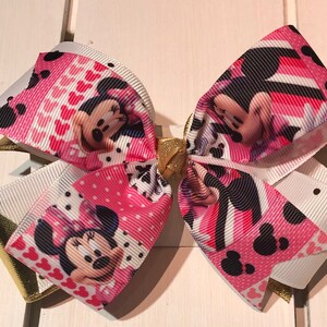 May include: A pink and white bow with a Minnie Mouse print. The bow has a gold accent in the center.