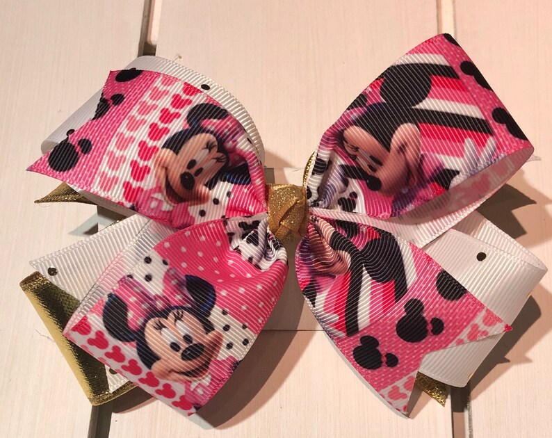 May include: A pink and white bow with a Minnie Mouse print. The bow has a gold accent in the center.
