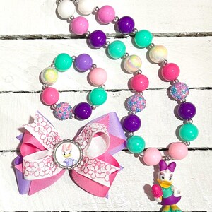 Pu&ograve; includere: Un colorato set di gioielli per bambini con collana, braccialetto e fiocco per capelli. La collana e il braccialetto hanno grandi perline rotonde rosa, viola, verdi e bianche. Il fiocco per capelli presenta il personaggio di Paperina.