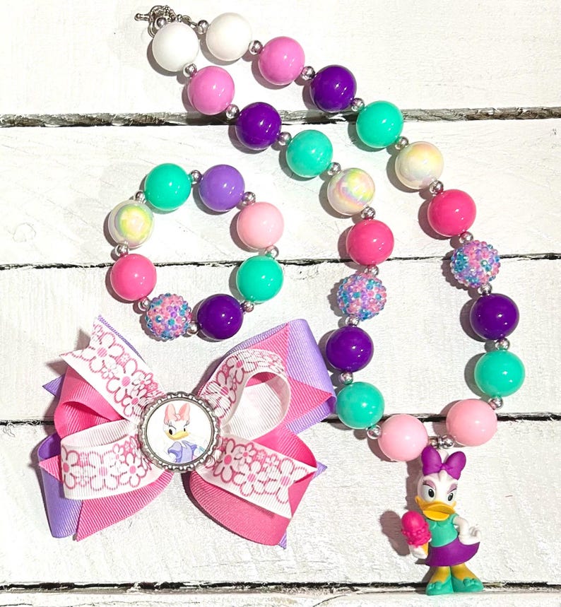 Pu&ograve; includere: Un colorato set di gioielli per bambini con collana, braccialetto e fiocco per capelli. La collana e il braccialetto hanno grandi perline rotonde rosa, viola, verdi e bianche. Il fiocco per capelli presenta il personaggio di Paperina.
