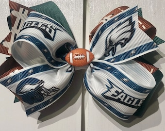 May include: A white, green, brown, and blue hair bow with a football design. The bow has the words "Eagles" and a football helmet on it.