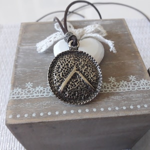 Peut inclure: Un collier avec un pendentif en forme de bouclier rond argenté, avec une surface texturée et un motif pointu en relief. Le collier est sur un cordon en cuir brun.