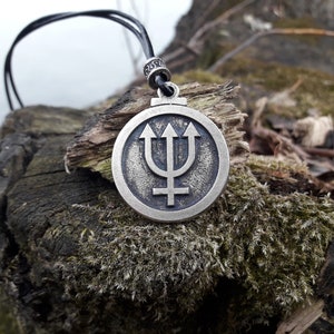 May include: A silver pendant necklace with a circular design featuring a trident and a symbol. The pendant is on a black cord and is resting on a mossy rock.