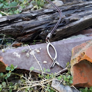 Puede incluir: Un collar con un colgante de Ichthys plateado en un cordón de cuero marrón. El collar está sobre una pila de ladrillos rotos y plantas verdes.
