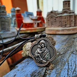 May include: A silver pendant necklace with a detailed image of the Virgin Mary holding a baby Jesus. The pendant is in the shape of a four-leaf clover and has a black leather cord.