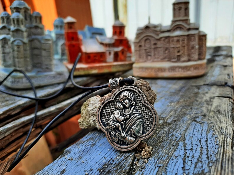 May include: A silver pendant necklace with a detailed image of the Virgin Mary holding a baby Jesus. The pendant is in the shape of a four-leaf clover and has a black leather cord.