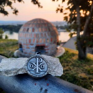 May include: A silver pendant with a Christian symbol, the Chi Rho, surrounded by a circle with the Greek letters Alpha and Omega. The pendant is resting on a piece of rock.