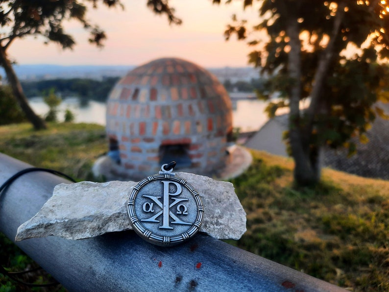 May include: A silver pendant with a Christian symbol, the Chi Rho, surrounded by a circle with the Greek letters Alpha and Omega. The pendant is resting on a piece of rock.