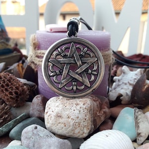 May include: A silver pendant necklace with a five-pointed star design. The star is surrounded by a decorative border. The necklace is hanging from a candle on a table with various decorative items.
