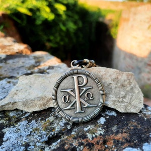 May include: A silver pendant with a Christian symbol, the Chi Rho, with the Greek letters alpha and omega on either side of the cross. The pendant is on a rock with a green background.
