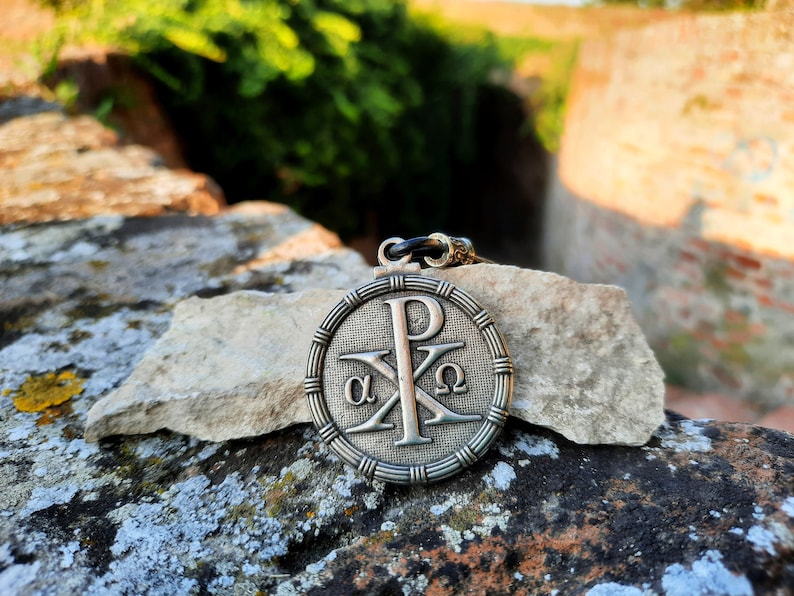 May include: A silver pendant with a Christian symbol, the Chi Rho, with the Greek letters alpha and omega on either side of the cross. The pendant is on a rock with a green background.