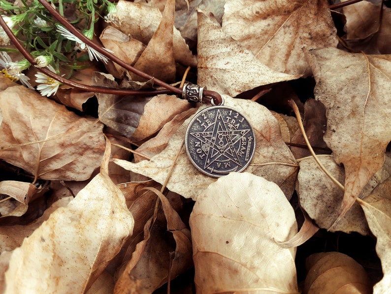May include: A silver pendant necklace with a five-pointed star design and inscription. The pendant is lying on a bed of brown leaves.