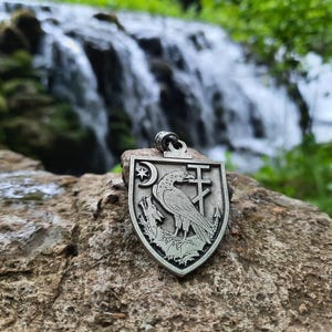 Puede incluir: Un colgante plateado con forma de escudo, con un cuervo, una luna creciente con una estrella y una cruz. El colgante se muestra sobre una roca texturizada con una cascada al fondo. El cuervo está posado en una rama.
