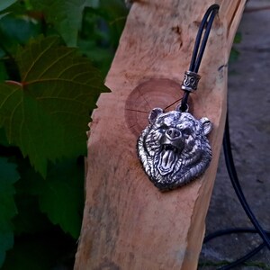 May include: A silver bear head pendant on a black leather cord. The pendant is detailed and shows the bear's teeth and tongue.