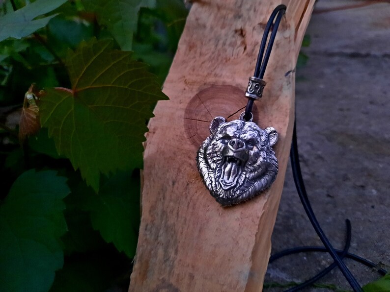 May include: A silver bear head pendant on a black leather cord. The pendant is detailed and shows the bear's teeth and tongue.
