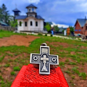 May include: A silver cross pendant with a circular design on each side. The cross is sitting on a red surface with a blurred background of a white building against a blue sky.