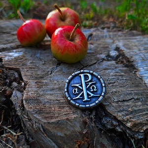 Könnte beinhalten: Eine silberfarbene Metallnadel mit den griechischen Buchstaben 'Alpha' und 'Omega' und dem Buchstaben 'P' in der Mitte, umgeben von einem Kreis aus kleinen Punkten. Die Nadel liegt auf einer Holzoberfläche mit vier roten Äpfeln.