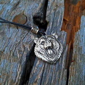 May include: A silver bear head pendant necklace with a black leather cord. The bear's mouth is open and its teeth are visible.
